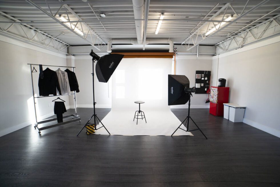Landscape shot showcasing photography studio set up for a photoshoot using white seamless paper backdrop and 2 strobes with model wardrobe hanging nearby.