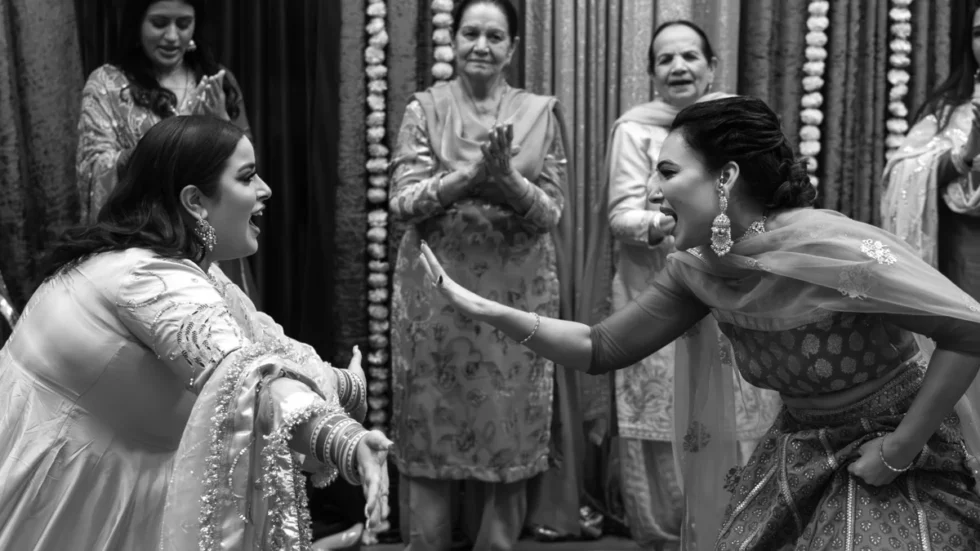 Two women performing a high-energy dance at an Indian celebration, surrounded by smiling family members